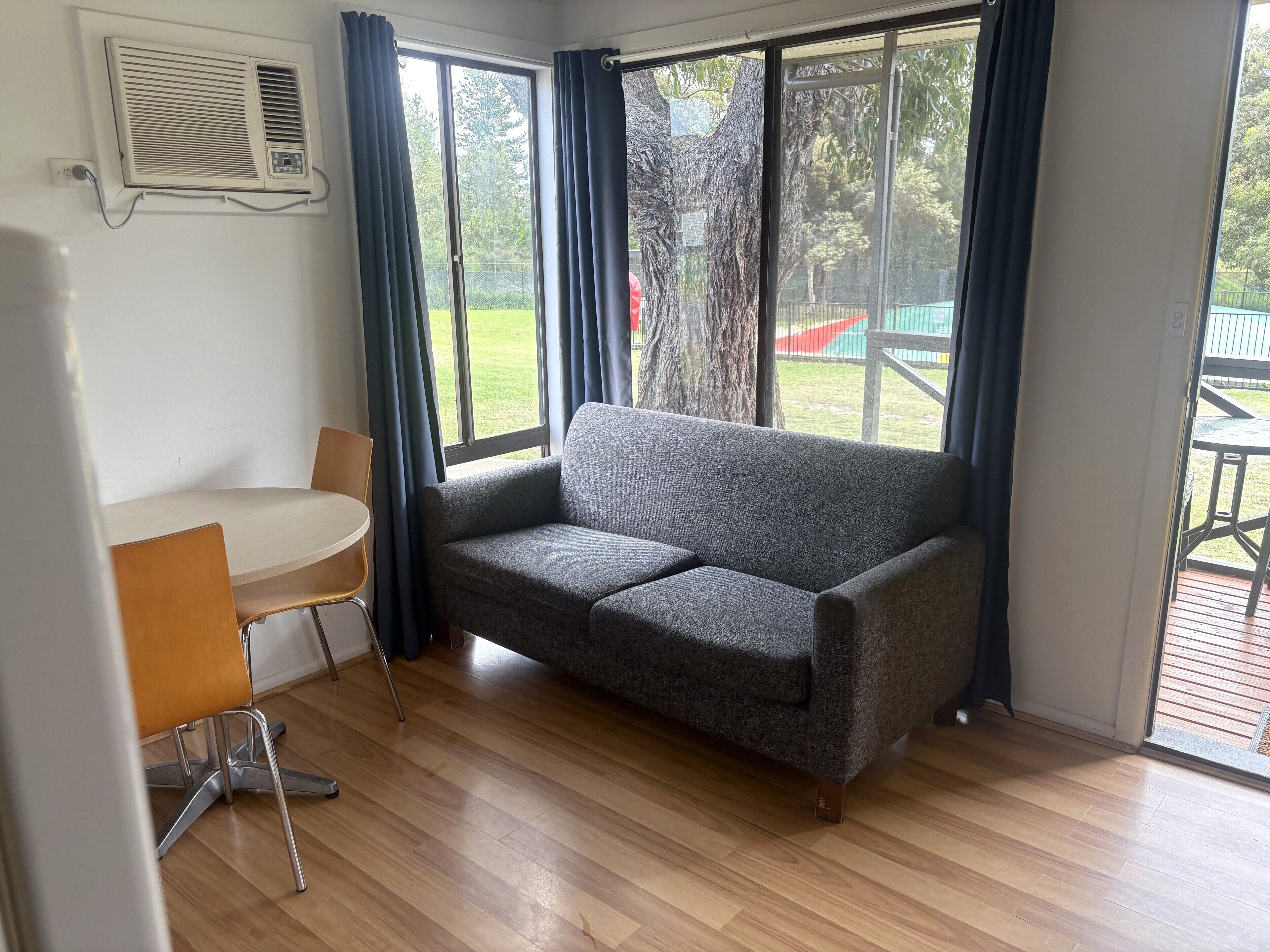 A room with a grey sofa, a small round table, and a chair. There is a large window with curtains, and a door leading outside.