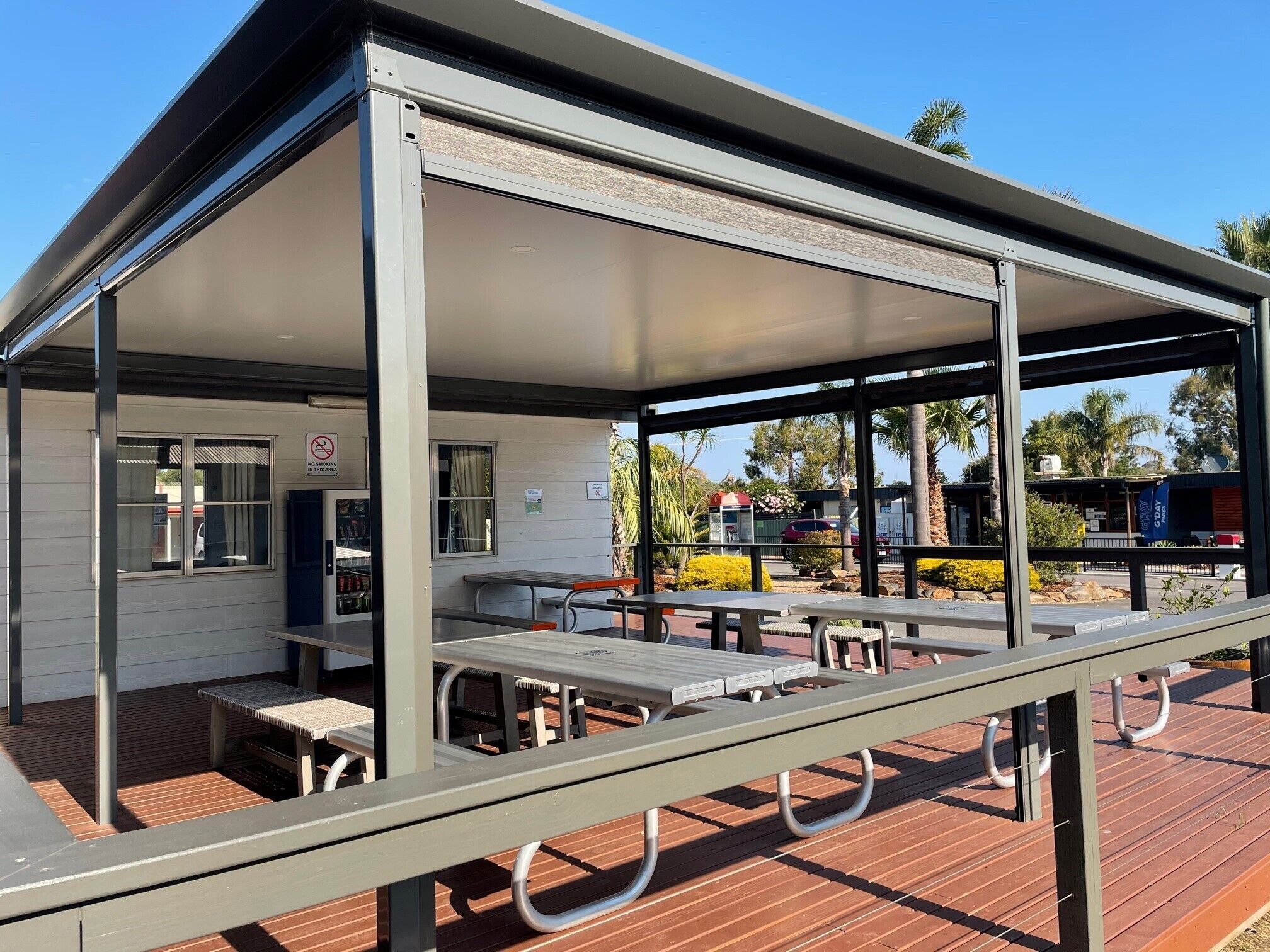 A covered picnic area with tables and benches, a building with a no-smoking sign, and a clear blue sky.
