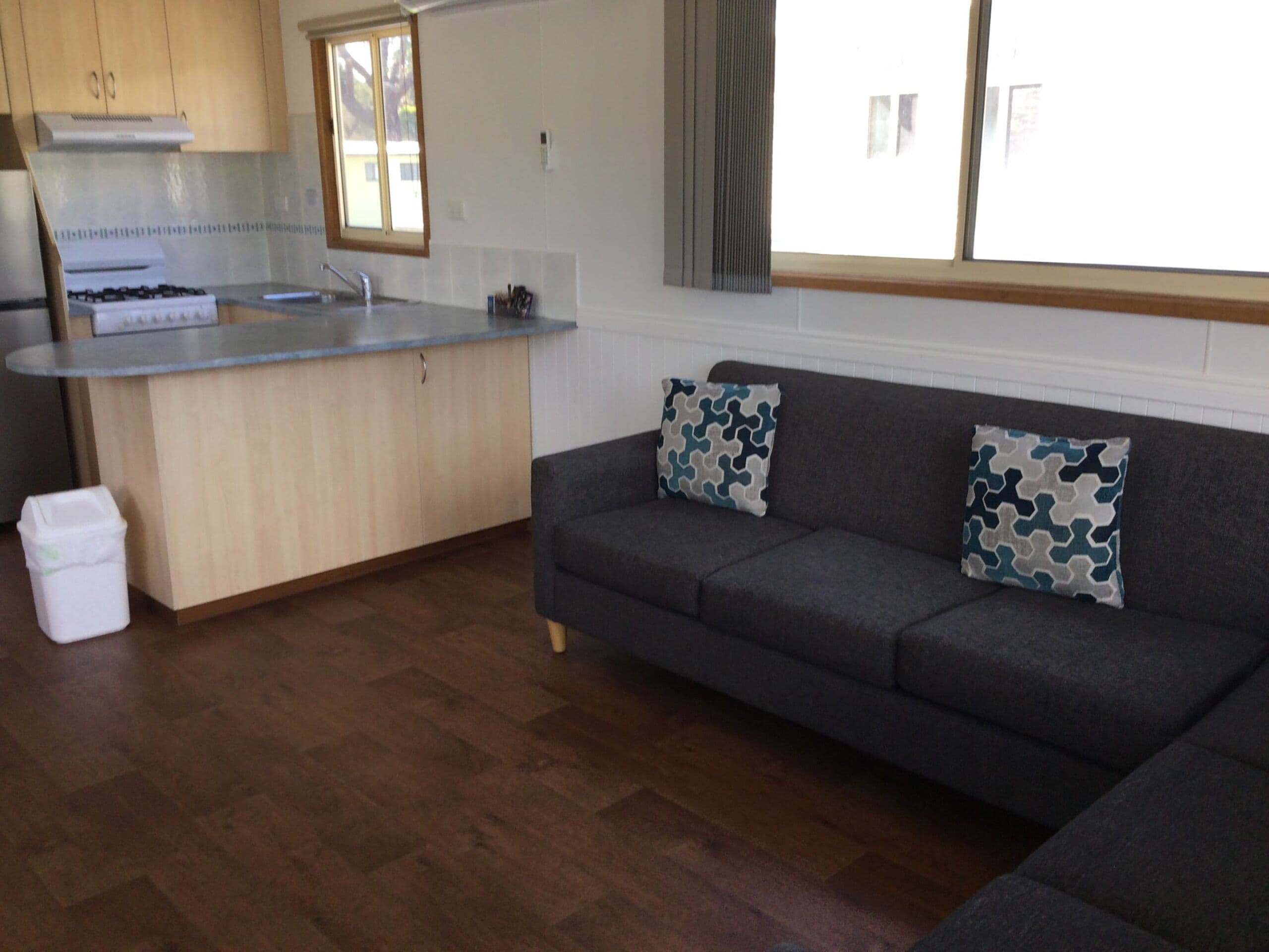 A kitchen with a built-in stove, a sink, and wooden cabinets. There is a grey sofa with patterned cushions and a white trash bin.
