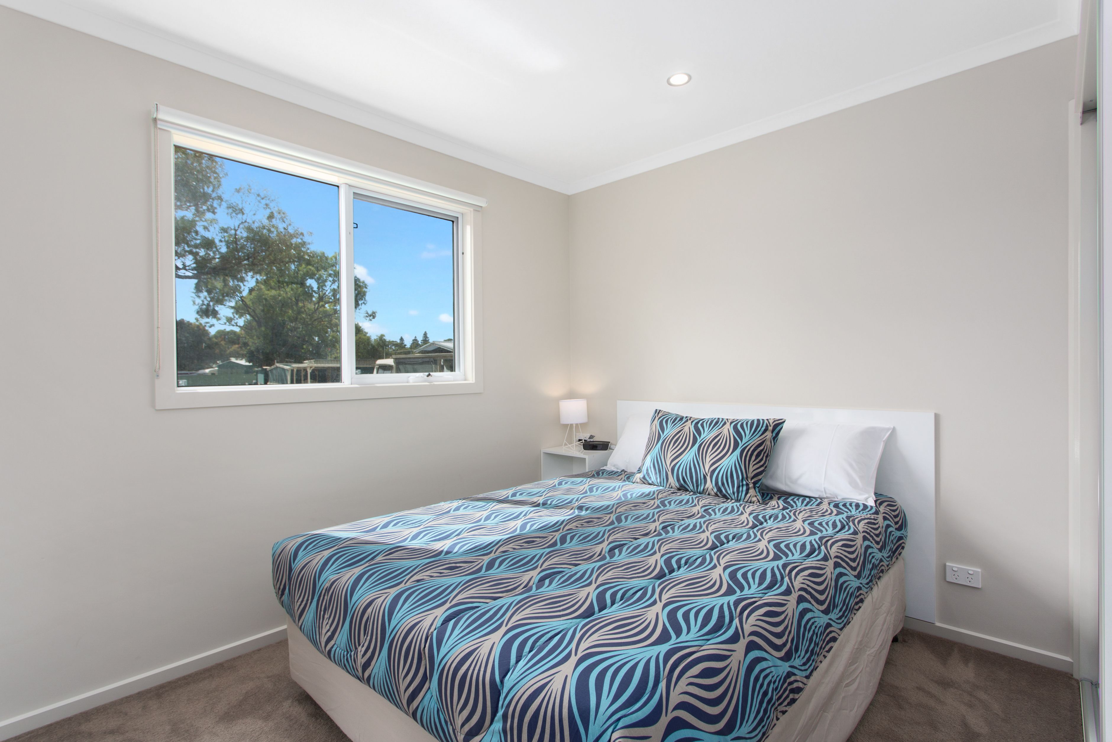 A bedroom with a large window, a bed with patterned bedding, a bedside table with a lamp, and a power outlet on the wall.