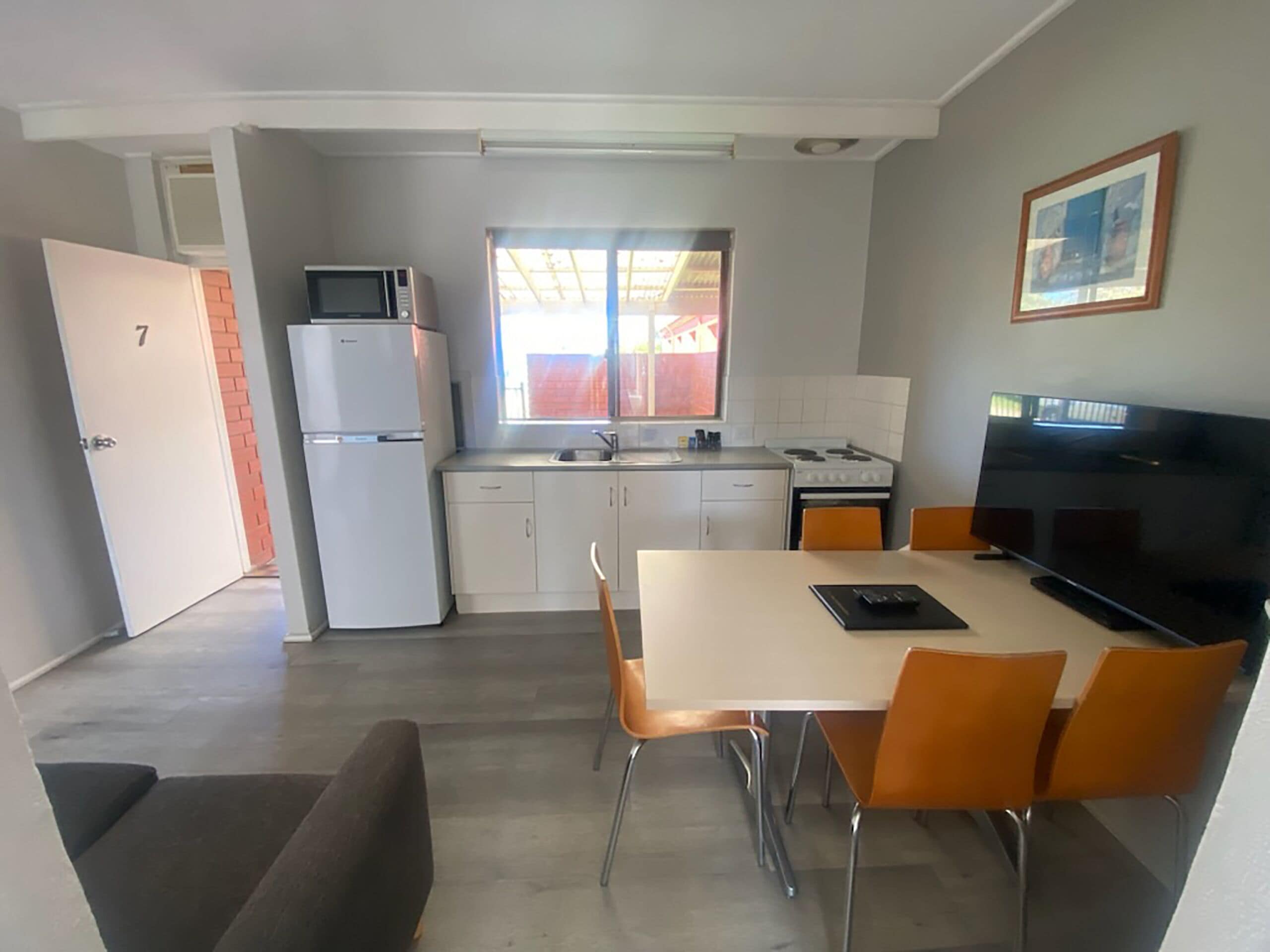 Cocina compacta equipada con refrigerador, microondas, estufa y lavamanos. Cuenta con una zona de comedor con mesa y sillas, además de un televisor sobre un soporte.
