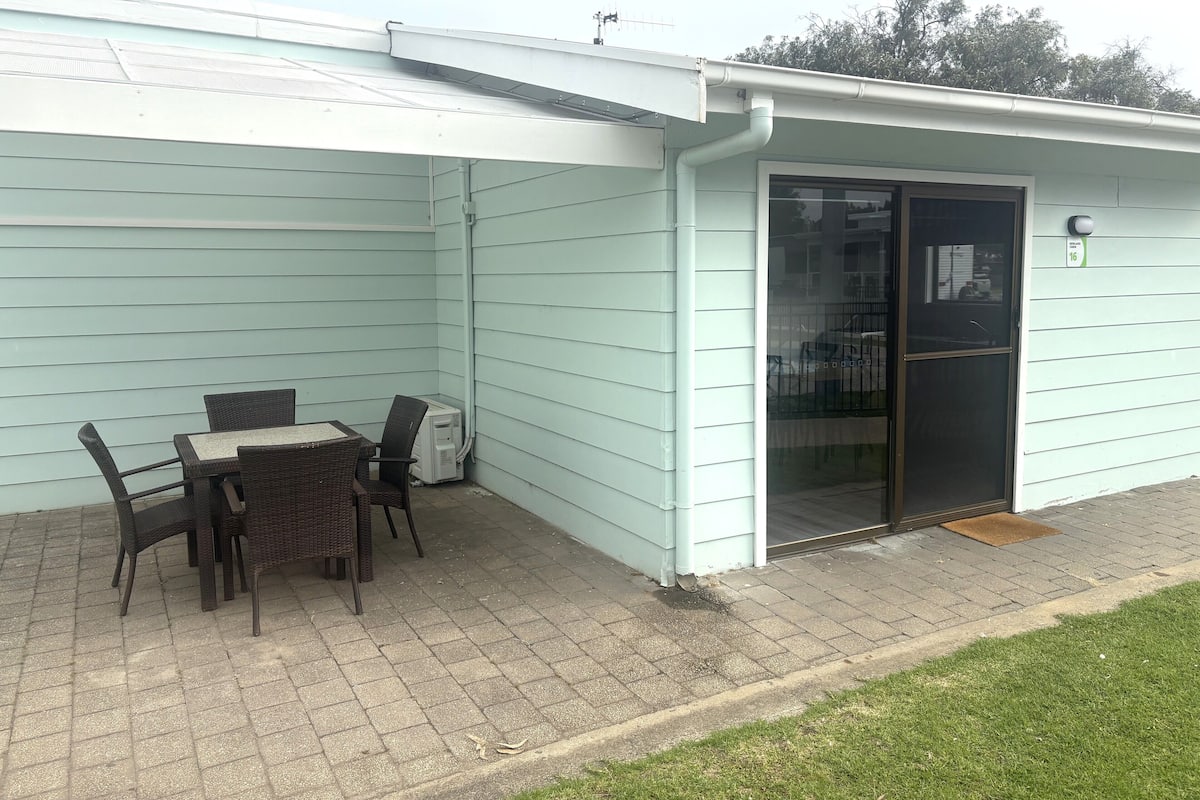 Een overdekt terras met een tafel en stoelen, een schuifpui en een kleine buitenruimte met een betegelde vloer en gazon.
