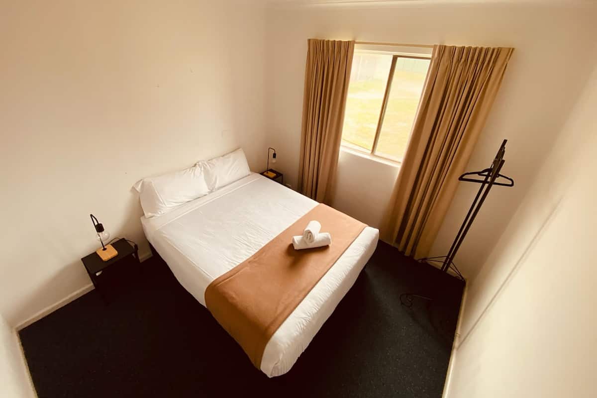 Een hotelkamer met een enkel bed, een nachtkastje met een lamp, een raam met gordijnen en een kapstok.