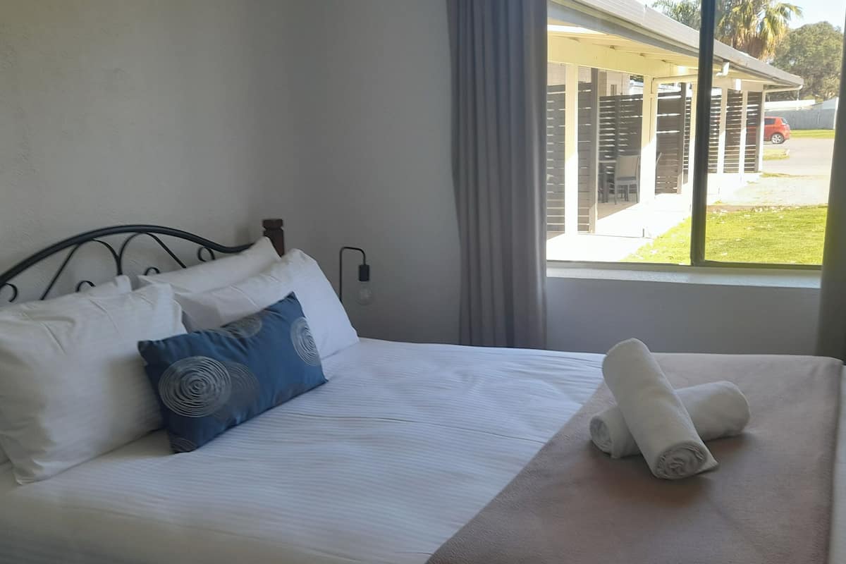 Een net opgemaakt bed met witte en blauwe kussens, een opgerolde handdoek, en een raam met uitzicht op een gebouw en een geparkeerde auto.