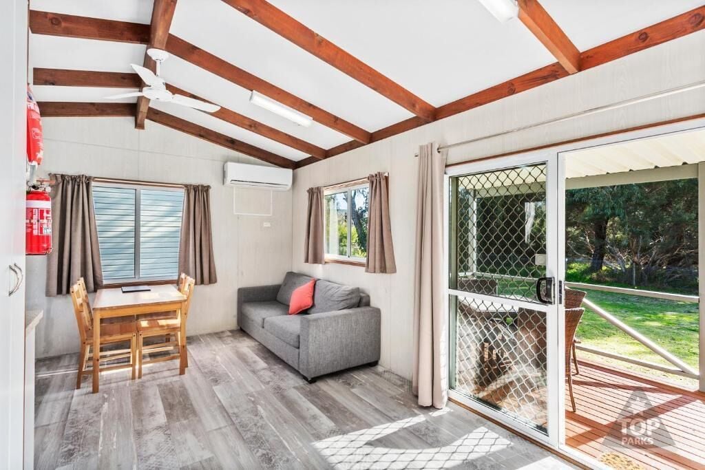 A room with a wooden ceiling, a grey sofa, a wooden table, and a sliding glass door to an outdoor area.