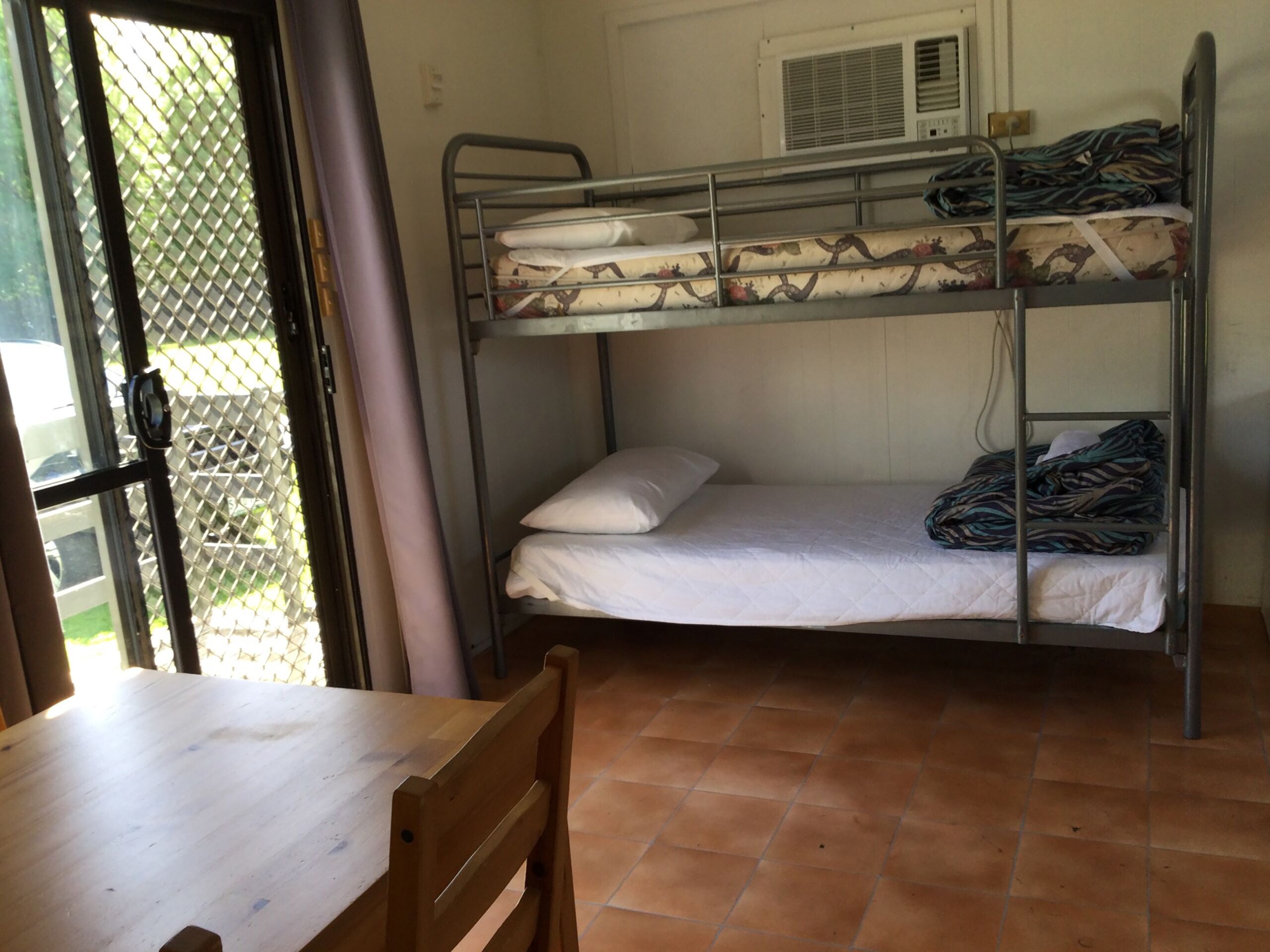 A bunk bed room with a table and chair, a window with a view of greenery, and an air conditioning unit on the wall.