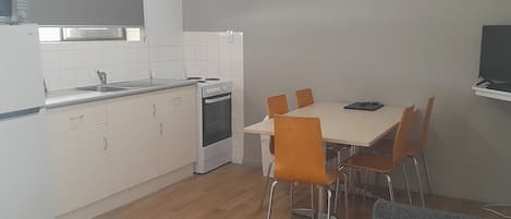 A compact kitchen with white cabinets, a stove, and a sink. There is a dining table with four chairs and a grey sofa in the foreground.