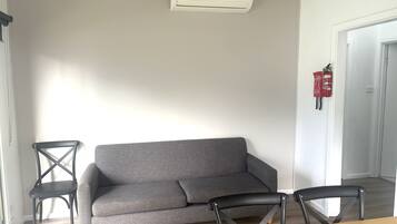 A living room with a grey sofa, two black chairs, a wooden table, and a white air conditioning unit on the wall.
