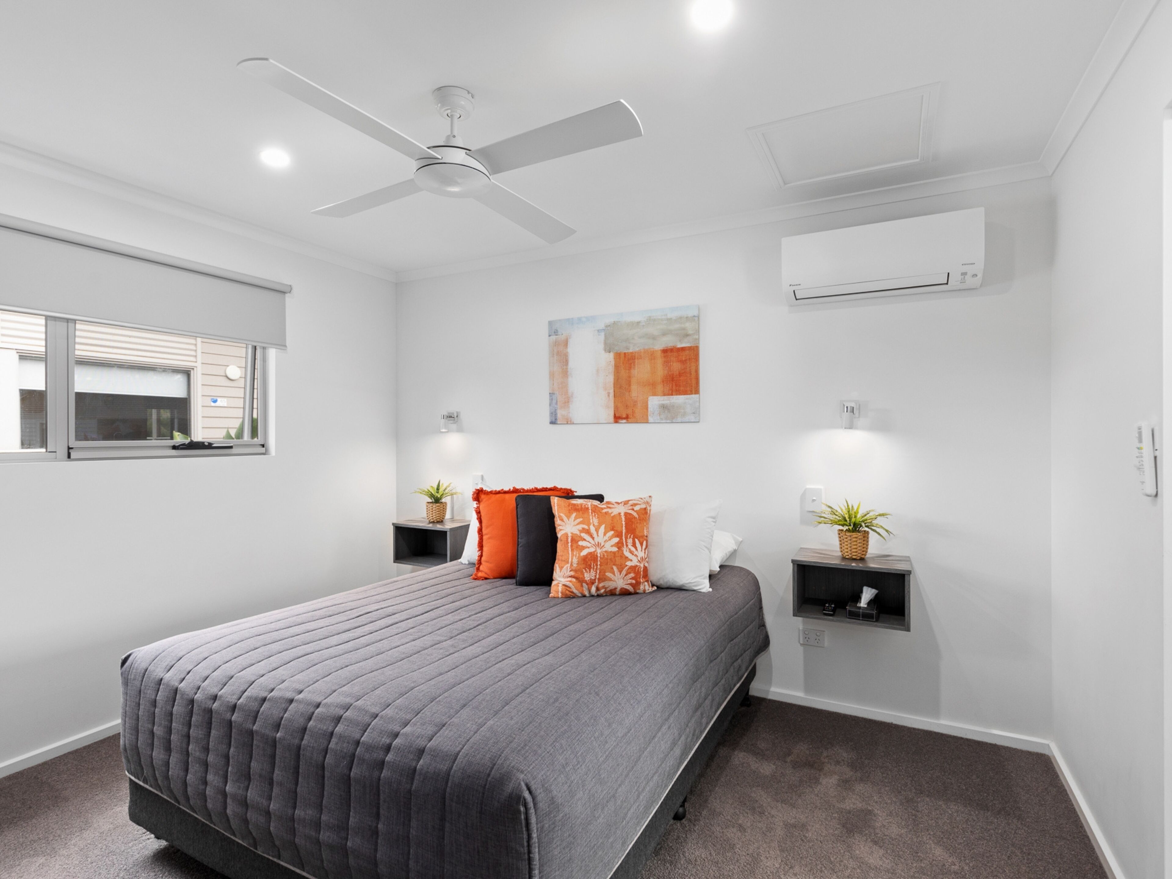 A bedroom with a bed, a ceiling fan, an air conditioning unit, a window, and a wall-mounted artwork.
