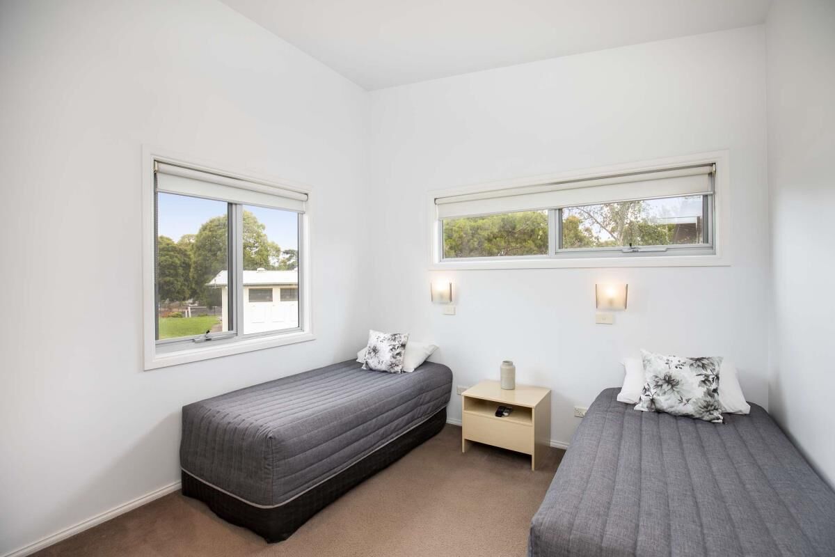 Une chambre avec deux lits simples, chacun muni d’un oreiller et d’une table de chevet. Il y a deux fenêtres, une sur chaque mur, et une petite table d’appoint entre les lits.