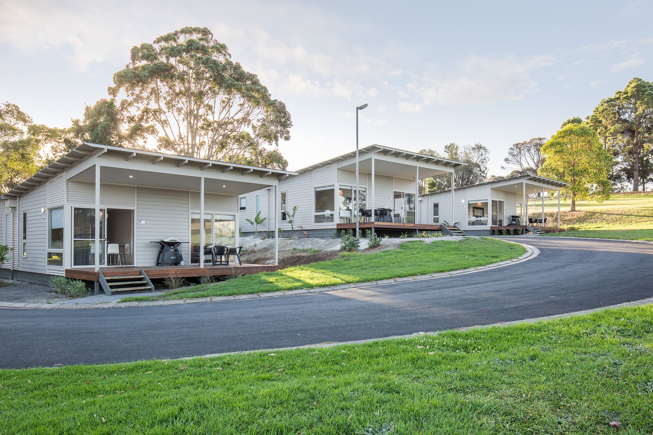 A row of two-story mobile homes with attached porches, a paved driveway, and surrounding greenery.