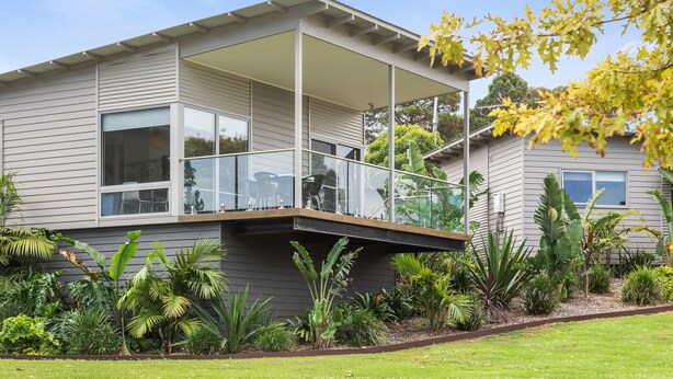 Une maison moderne à deux étages, dotée d’un balcon couvert, de grandes fenêtres et d’un jardin bien entretenu où poussent différentes espèces de plantes.