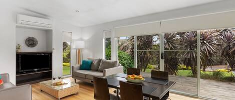 A modern living room with a flat-screen TV, a sofa, a dining table with chairs, and a ceiling fan.