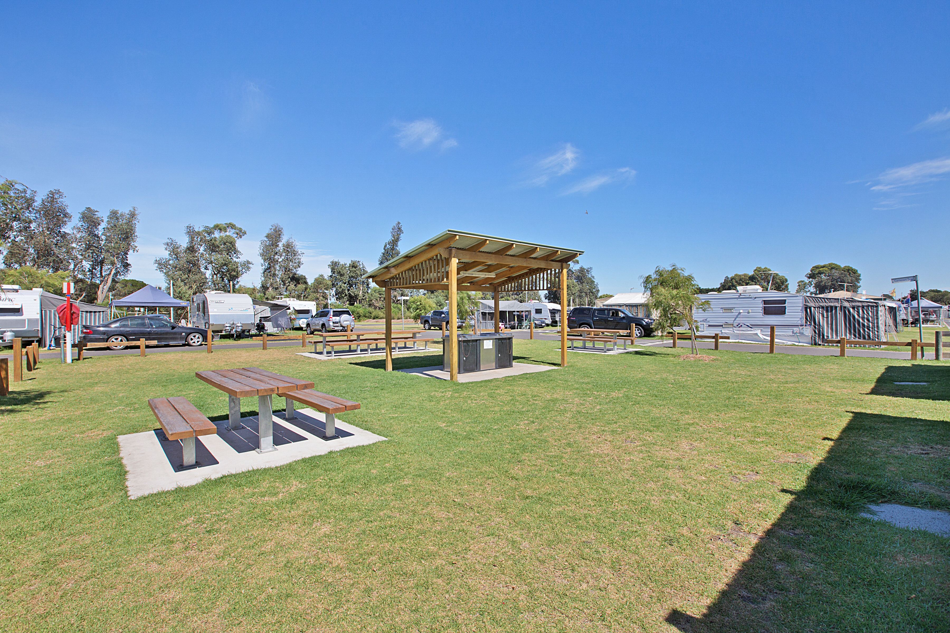 A campground with picnic tables, a covered pavilion, and RVs parked in the background.