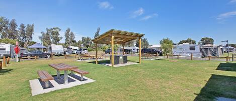 A campground with picnic tables, a covered pavilion, and RVs parked in the background.