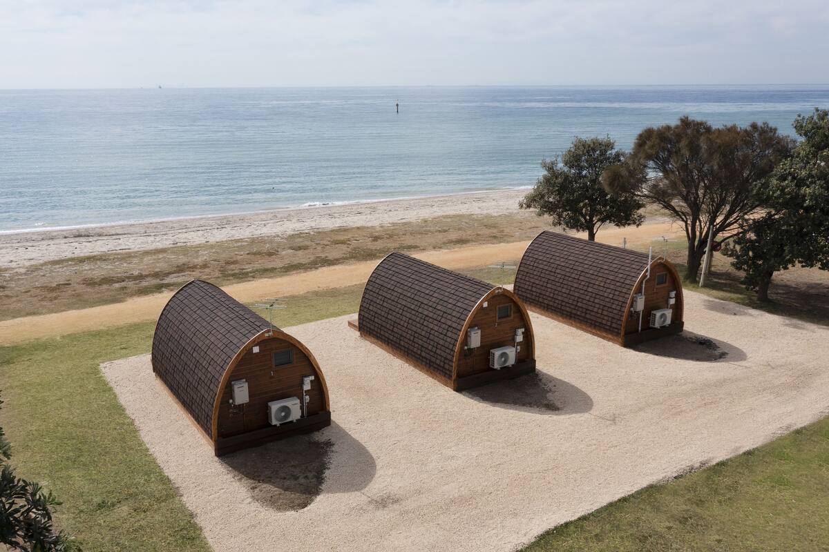 Drie houten, gebogen, overdekte constructies met airconditioners langs een kiezelpad aan het strand.