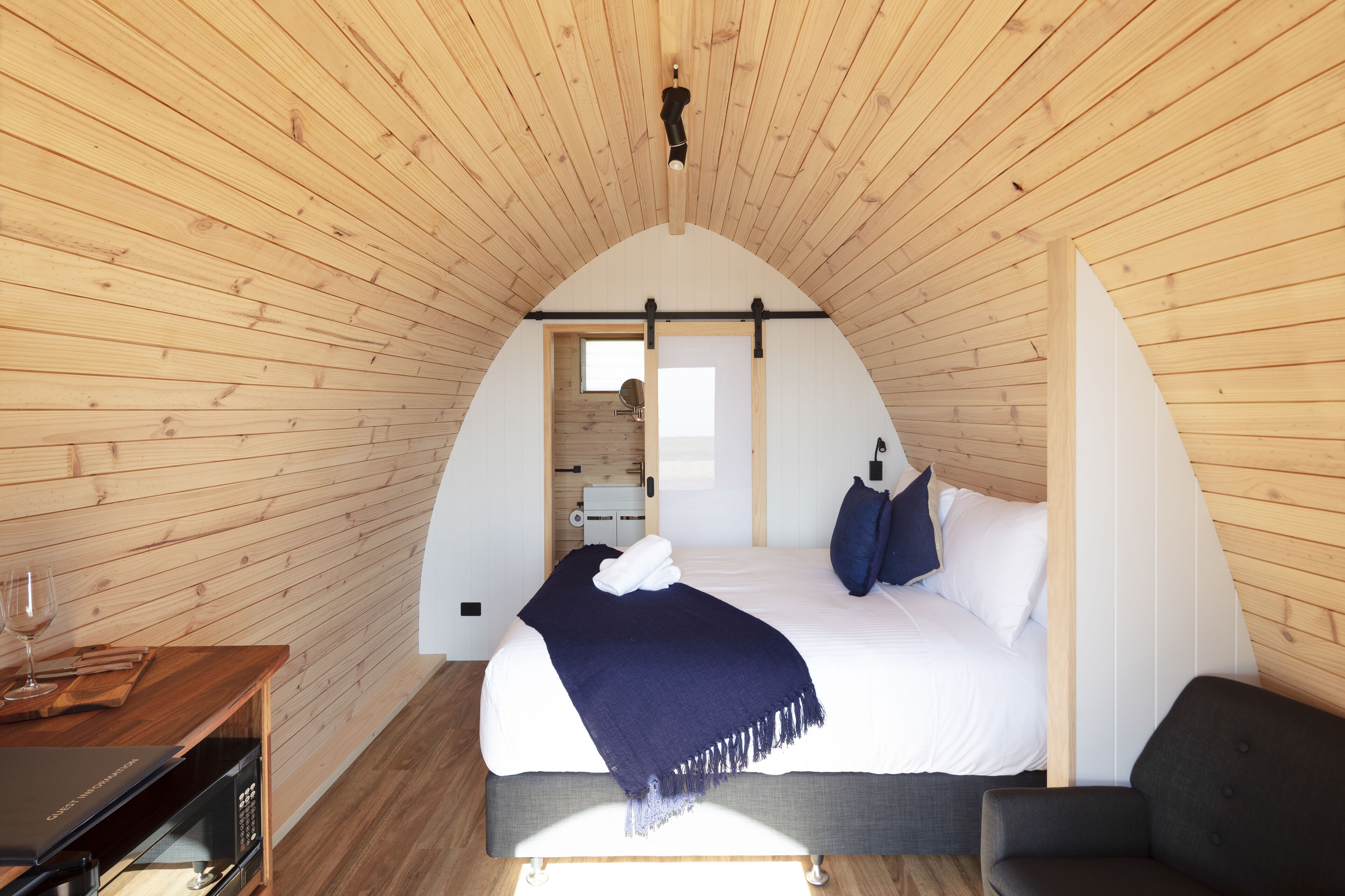 Ein Schlafzimmer mit Holzbalkendecke, einem Bett mit weißen Leinen, einer dunkelblauen Tagesdecke und einem kleinen Tisch mit einem Glas.