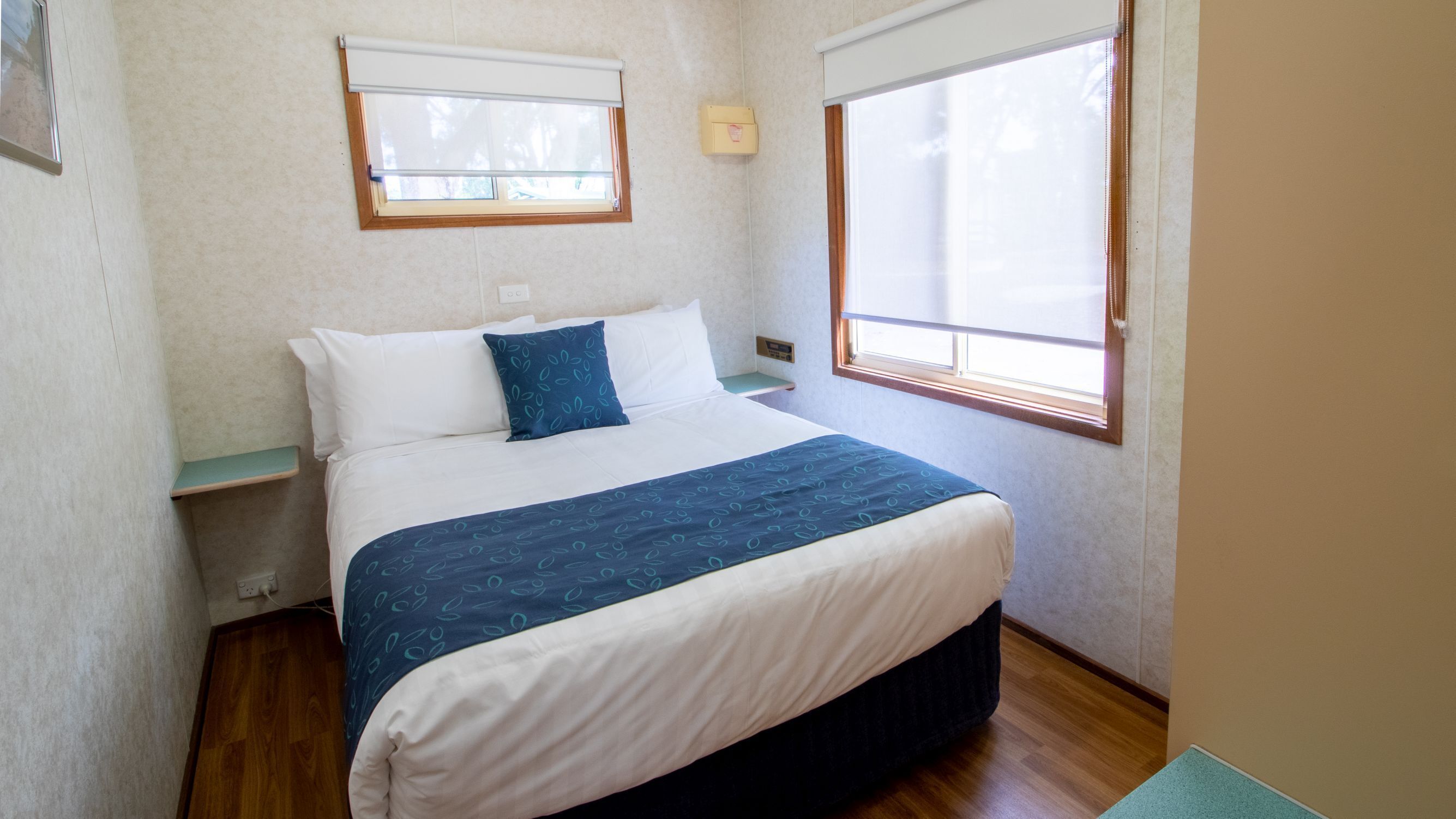 A bedroom with a bed, two bedside tables, a window with blinds, and a wall-mounted light switch.