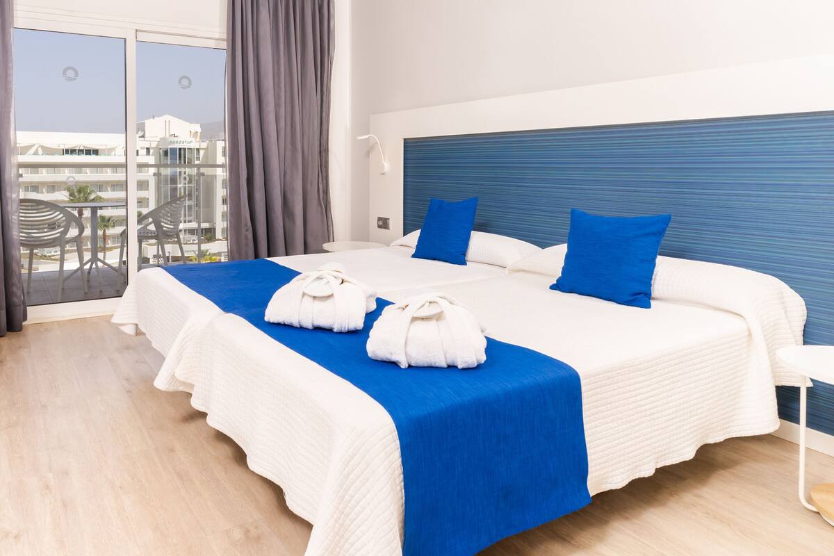 A hotel room with a large bed, a view of the city, and a balcony with chairs.