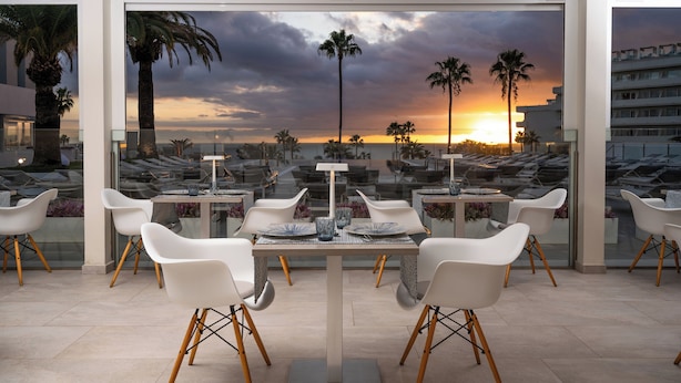 A modern outdoor dining area with a glass table and white chairs, overlooking a sunset view with palm trees and a building in the background. - HOVIMA Cool Costa Adeje (Adeje)