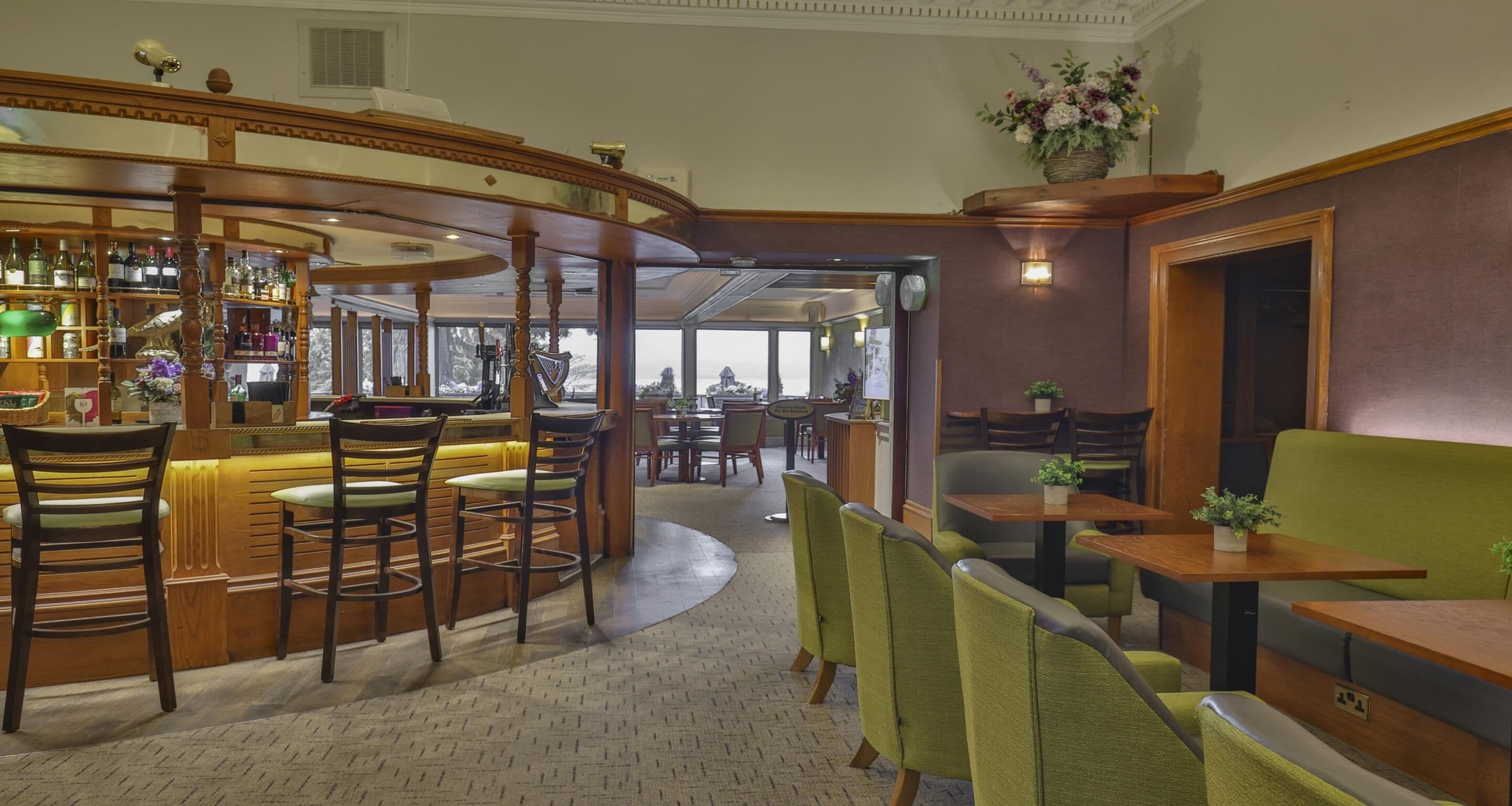 A bar area with wooden bar stools and a variety of bottles. There are tables and chairs for dining, and a green sofa.