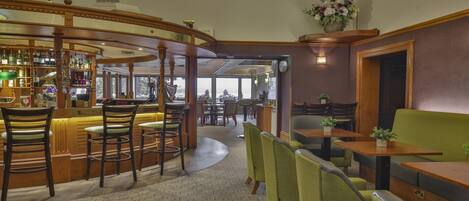 A bar area with wooden bar stools and a variety of bottles. There are tables and chairs for dining, and a green sofa.