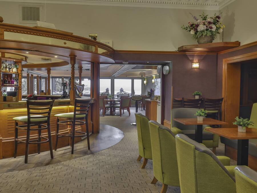 Et område med bar, træbarguide og et udvalg af flasker. Der er borde og stole til spisning, samt en grøn sofa.