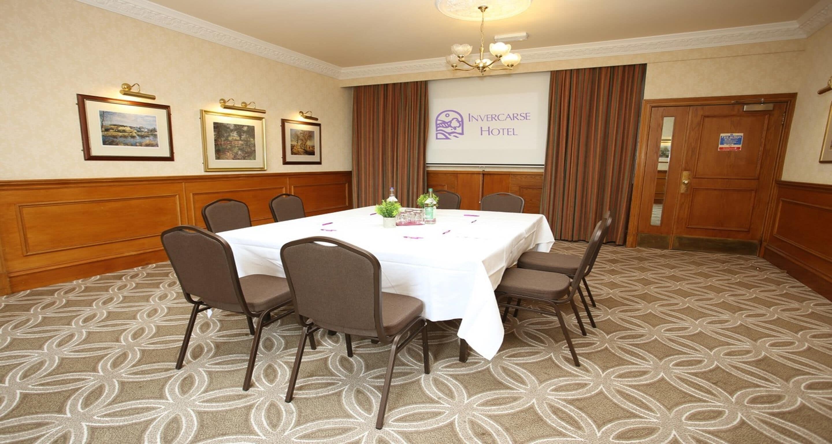 A conference room with a white table, brown chairs, framed pictures on the wall, and a projector screen displaying the hotel name.