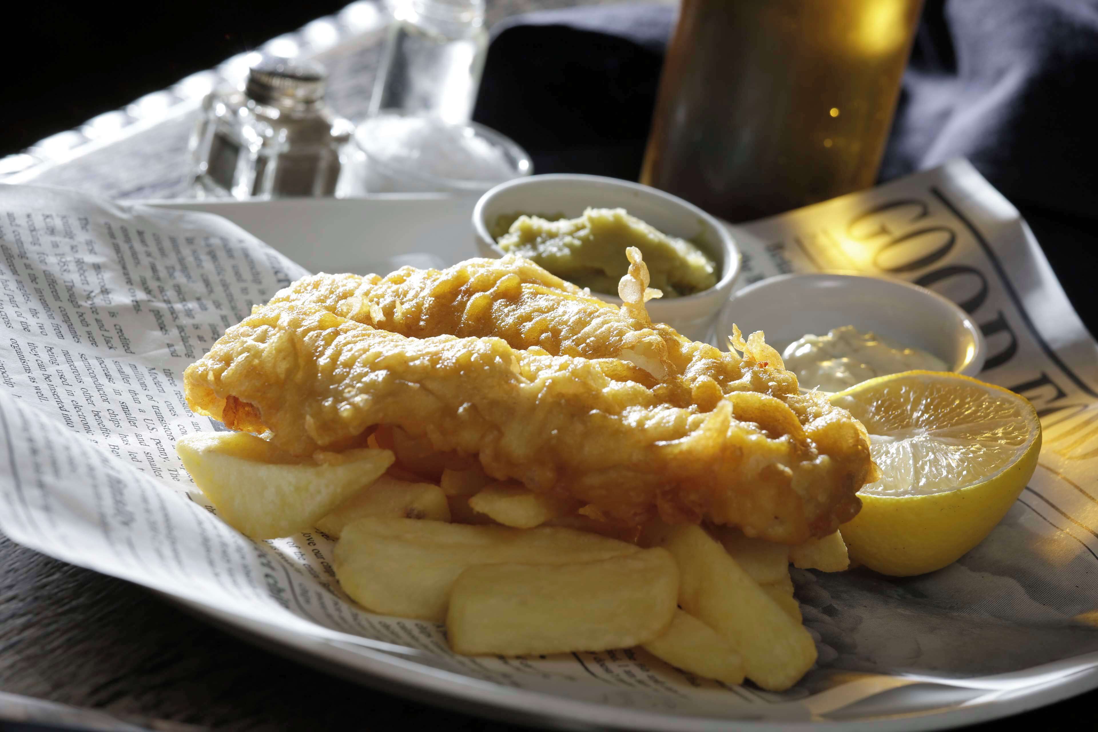 Ein Teller Fish and Chips mit Zitronenspalten und Erbsenpüree, serviert auf Zeitungspapier.
