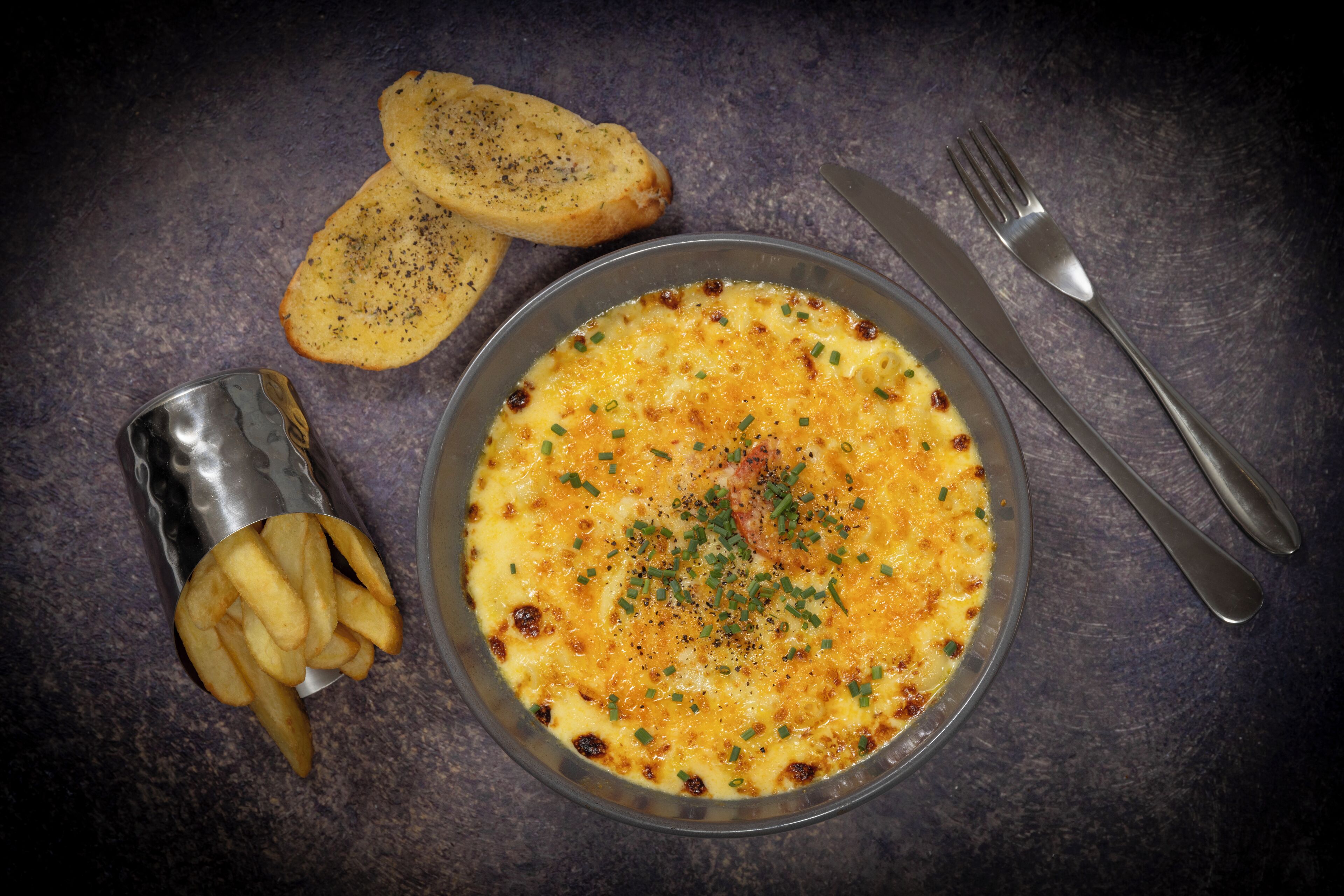 A plate of food with a side of fries and bread, accompanied by cutlery on a dark surface.