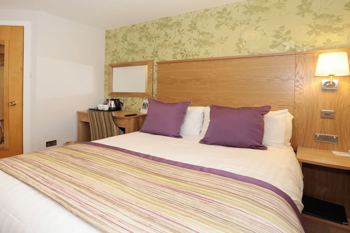 A hotel room with a bed, a wooden headboard, a desk with a chair, a mirror, and a nightstand with a lamp.