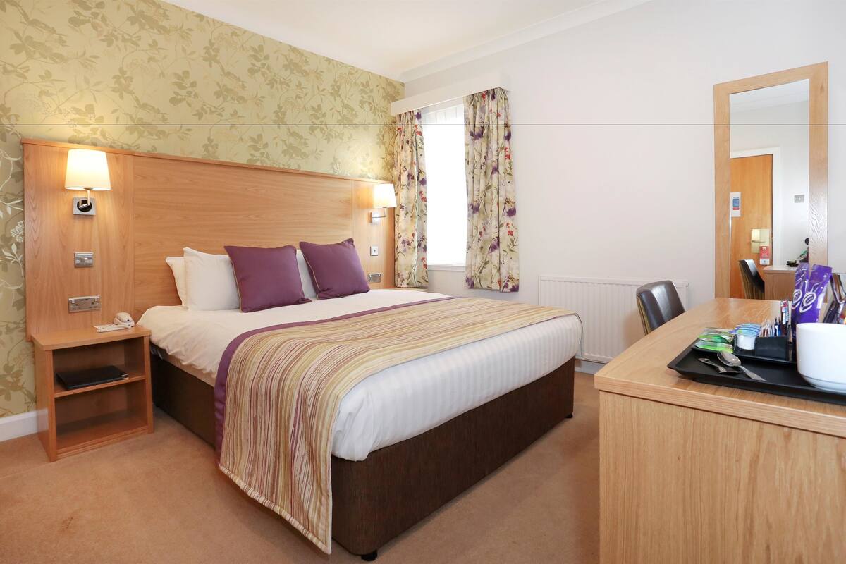 A hotel room with a wooden headboard, a bed with a striped blanket, a nightstand with a lamp, a desk with a tray of toiletries, and a floral curtain.
