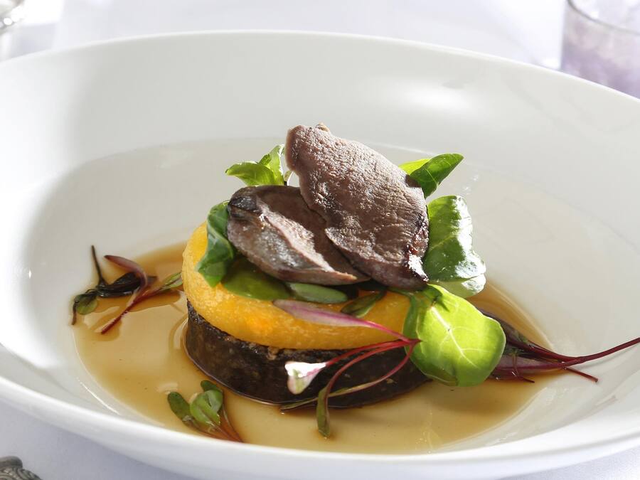Un tazón blanco con un plato que contiene carne, verduras y un elemento amarillo, posiblemente una fruta o verdura.