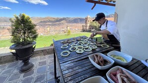 A chef preparing food on an outdoor grill with a scenic view of a city and mountains in the background. - Palacio Manco Capac by Ananay Hotels (Cusco)