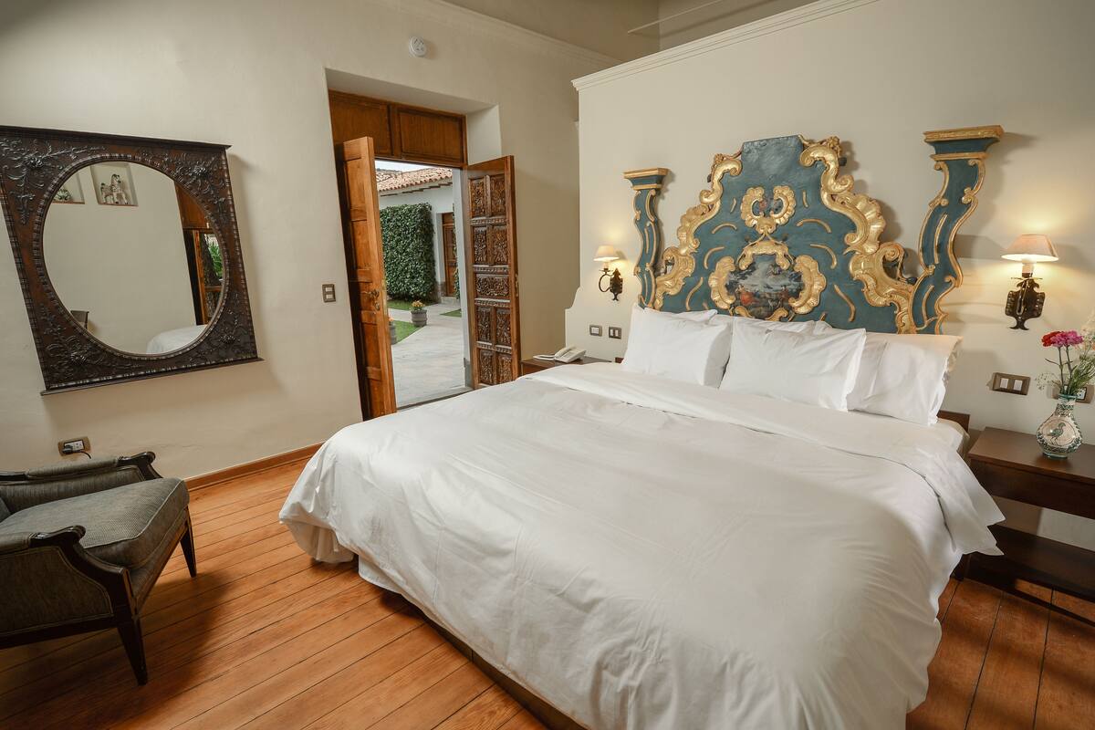 Un dormitorio amplio con una cama grande, piso de madera, una silla, un espejo y un cabecero decorativo.