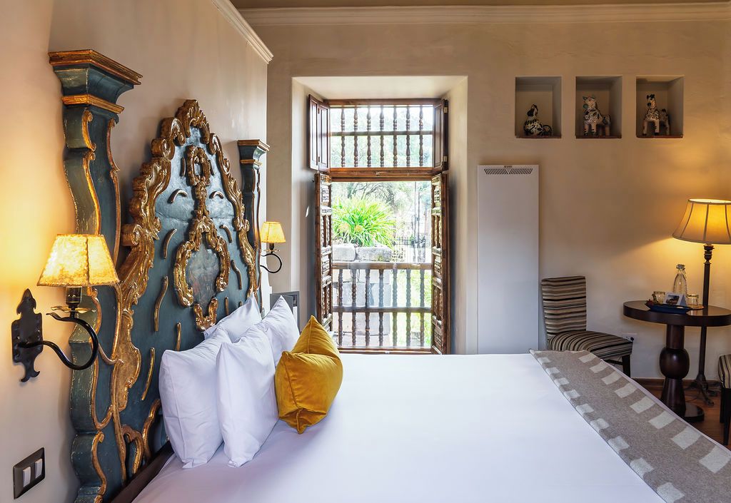 A hotel room with a large bed, ornate headboard, two wall-mounted lamps, a small table with a lamp, and a window with a view of greenery.