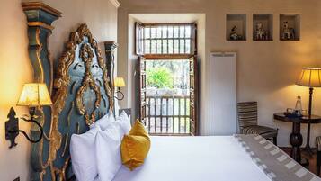 A hotel room with a large bed, ornate headboard, two wall-mounted lamps, a small table with a lamp, and a window with a view of greenery.