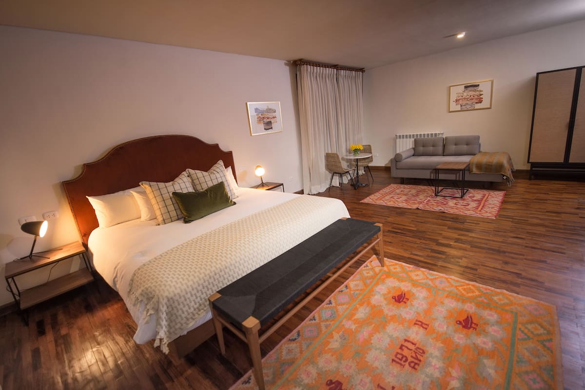 Un dormitorio con una cama grande, mesitas de noche, un banco, un sofá, una silla y una pequeña mesa con un jarrón.