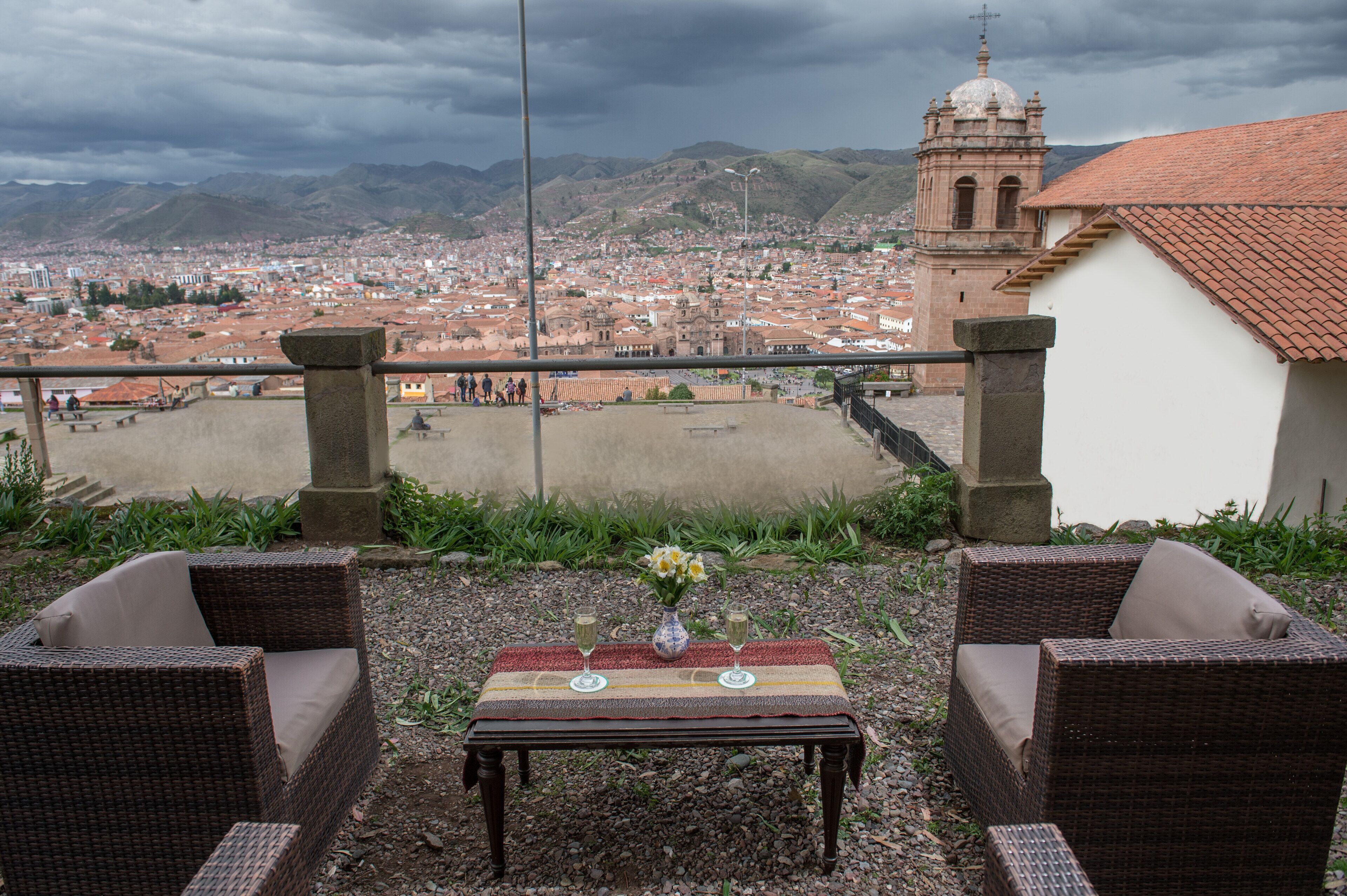 A wicker chair and table set with a vase and two glasses, overlooking a cityscape with a prominent bell tower.
