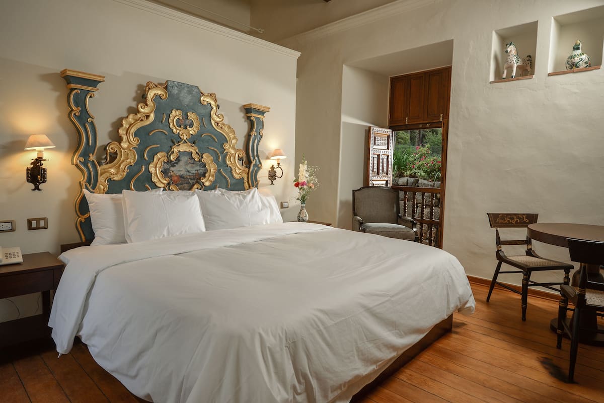 Una cama grande con cabecera decorada, una mesita de noche de madera con lámpara, una silla y una pequeña mesa en una habitación con suelo de madera y vistas a zonas verdes a través de una ventana.