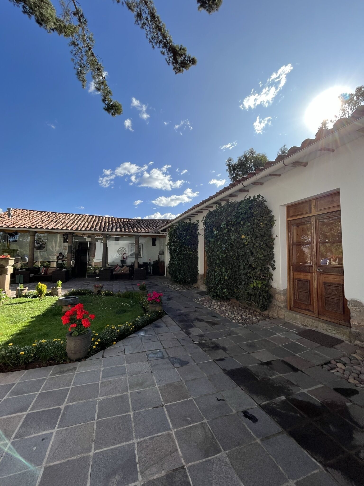 Un patio con suelo de baldosas, un jardín con plantas en macetas y un edificio con tejado de tejas y puerta de madera.