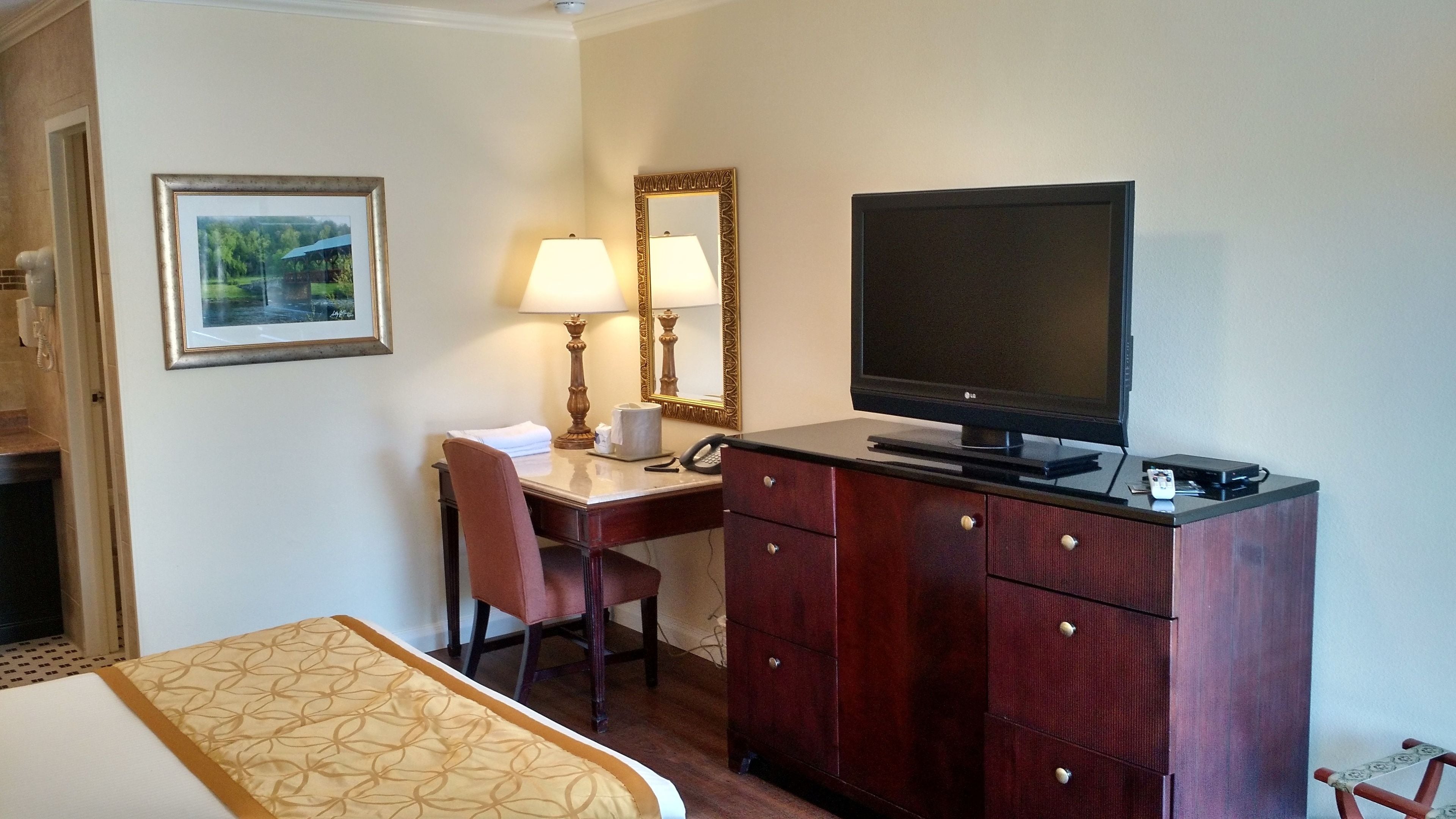 A hotel room with a bed, a desk with a chair, a television, a dresser, and a framed picture on the wall.