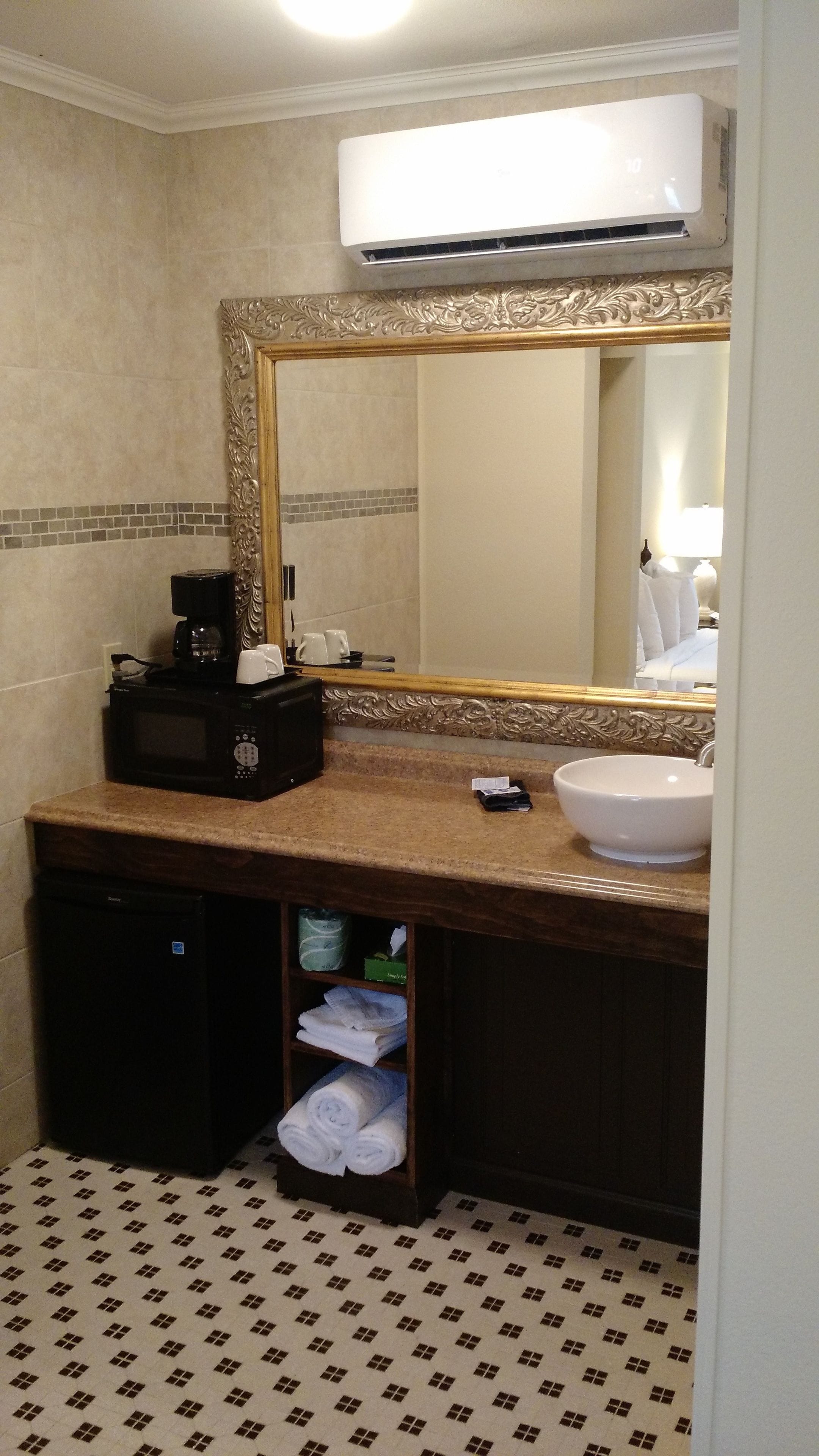 A bathroom with a vanity, sink, and mirror. There is a microwave, a coffee maker, and a towel rack.