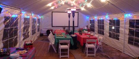 A tented dining area with tables set for a meal, string lights, and a projector screen.