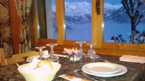 Una mesa de comedor dispuesta con platos, vasos y cubiertos, con vistas a montañas nevadas a través de una ventana.