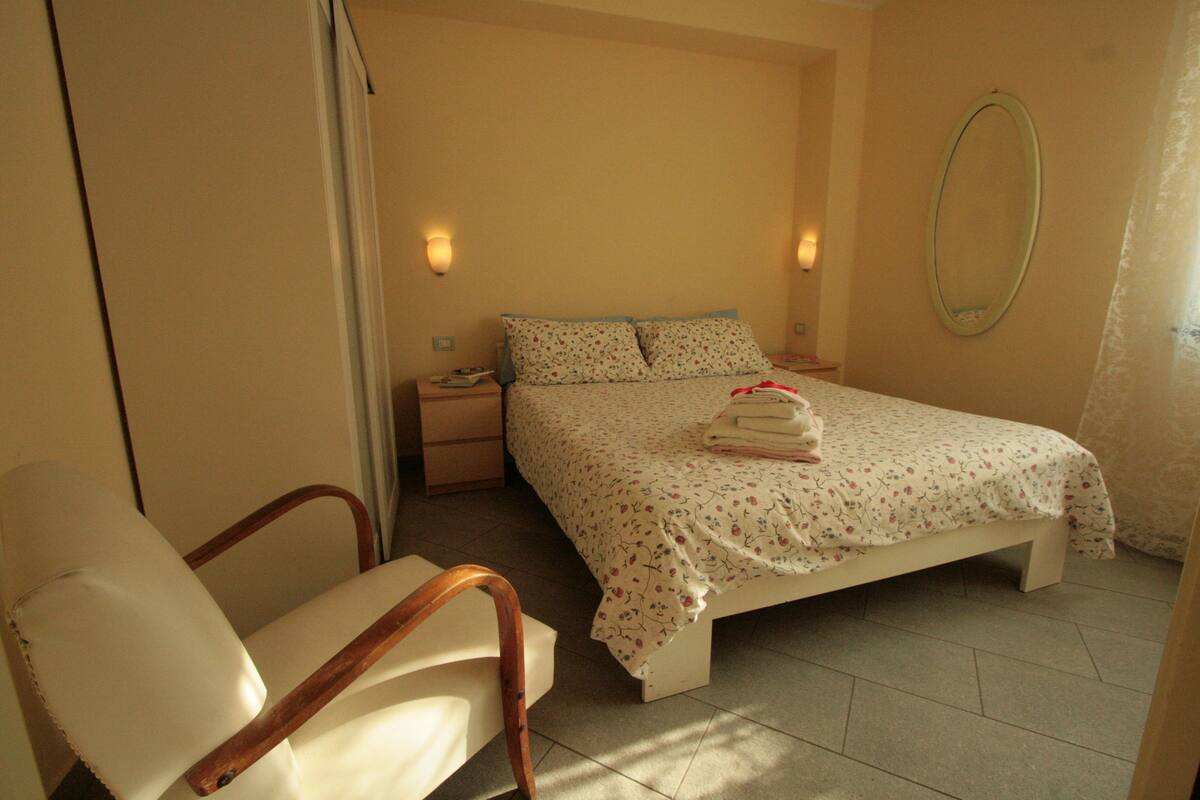 A bedroom with a bed, a chair, a nightstand, and a wall-mounted mirror.