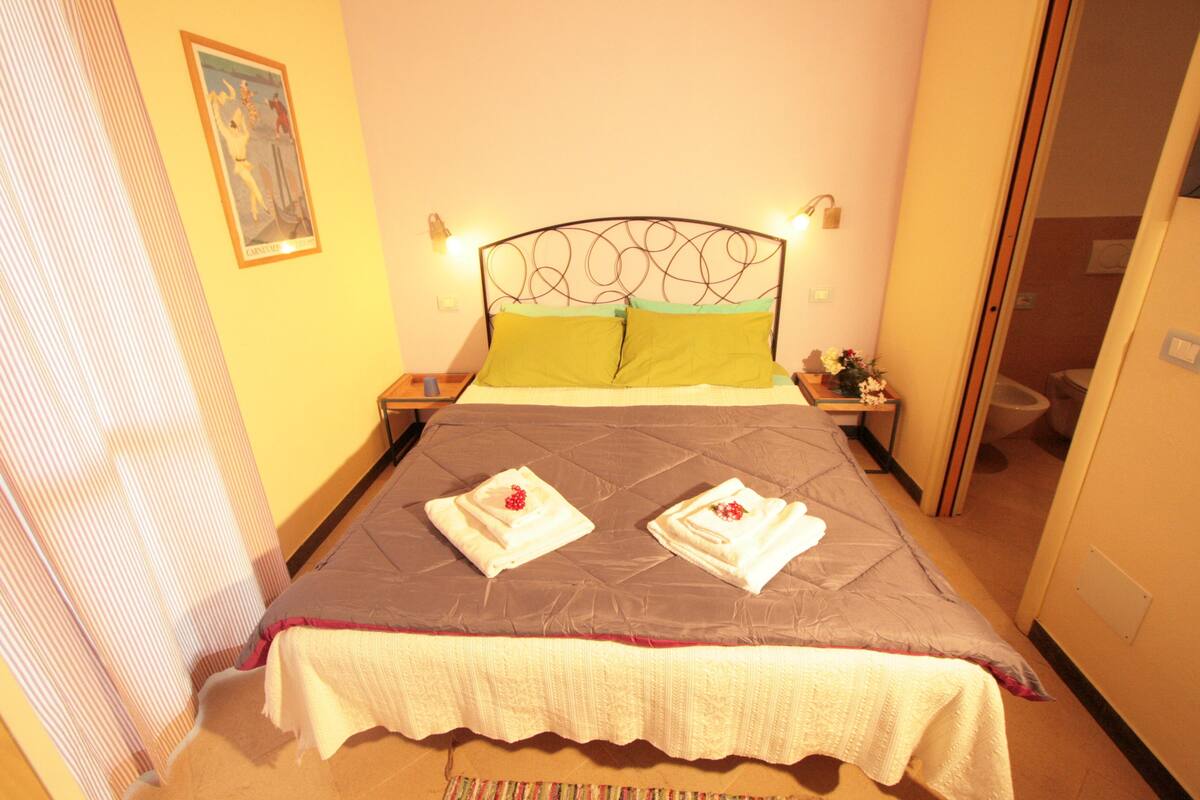 A neatly made bed with a quilted headboard, two pillows, and two towels with flowers. There is a nightstand with a lamp and a small table with a vase of flowers.