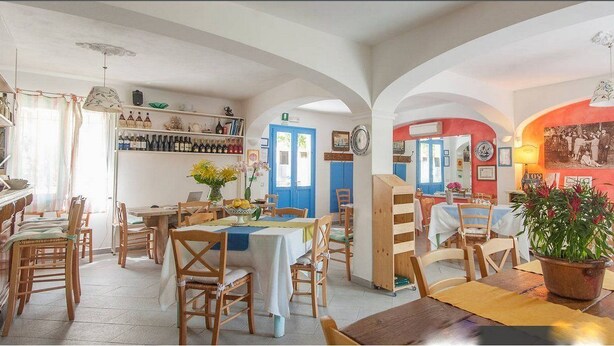Un comedor con sillas y mesas de madera, una cocina con estantes llenos de botellas, y una pared roja decorada con una foto familiar.