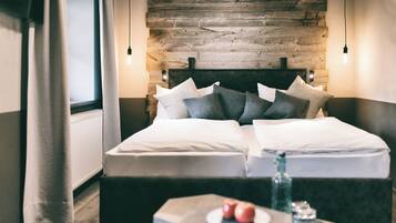 Una habitación de hotel moderna con cabecera de madera, dos camas con ropa blanca, una mesita de noche con una fuente de manzanas y un vaso, y vistas a la ventana con cortinas.