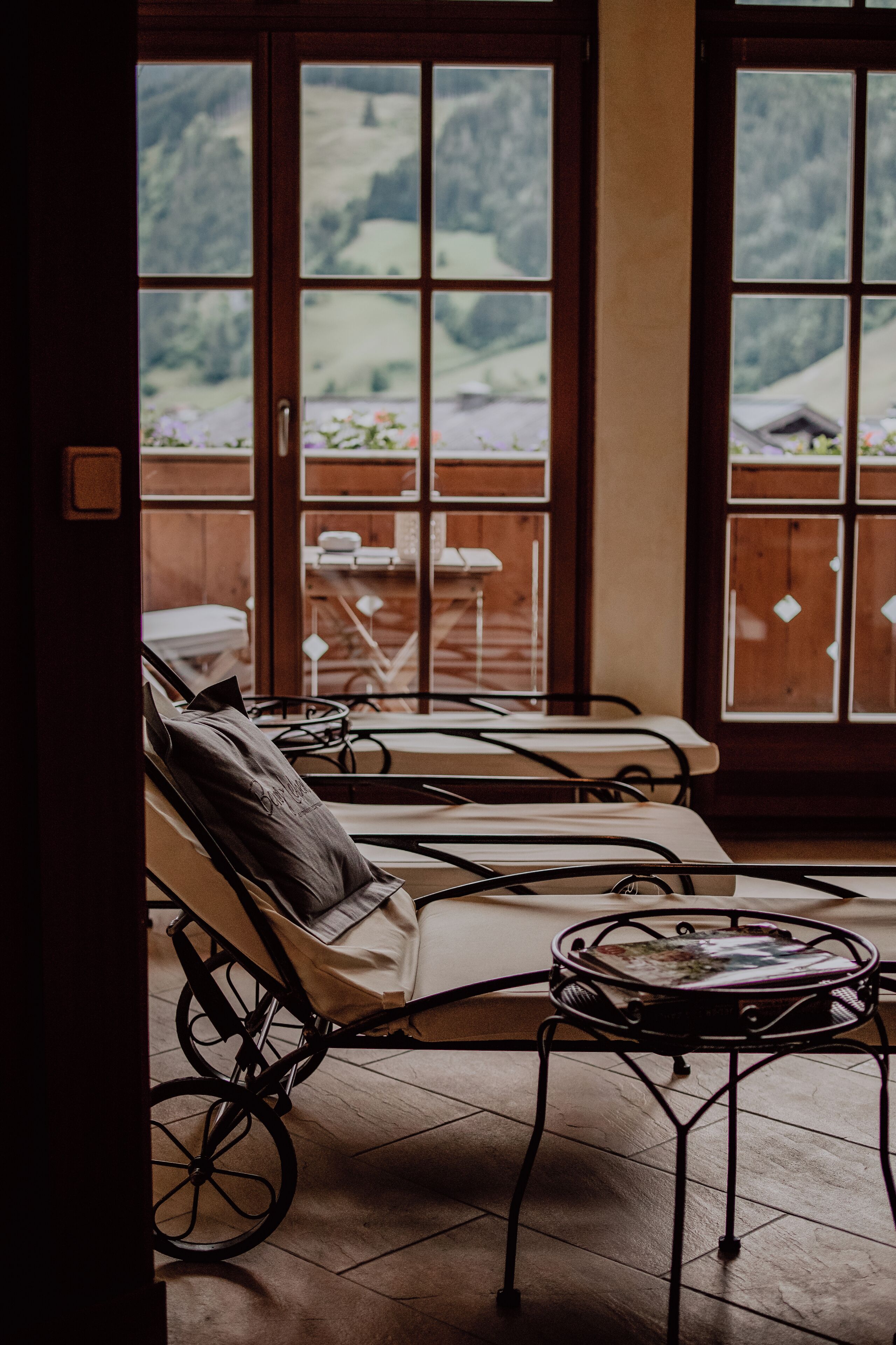A room with two sun loungers, a small table, and a view of a mountainous landscape through large windows.